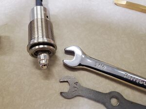 A stainless steel stem from the 4BSV on its side with a silver gasket sticking out of the top, next to it on the table are two different size wrenches, one at 1/4 inch and the other at 5/16 inch.